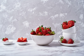 Ripe organic strawberries in white ceramic bowl and chamomile in vase on gray concrete background, copy space. Healthy food concept, still life