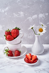Ripe organic strawberries in white ceramic bowl and chamomile in vase on gray concrete background, copy space. Healthy food concept, still life
