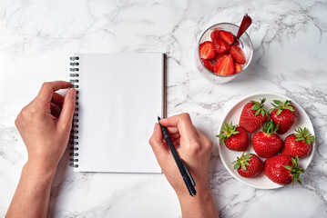 Top view of woman hands writing in blank notebook and glass of healthy yogurt with fresh sliced strawberry on white table, copy space. Healthy food concept. Flat lay