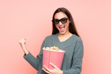 Young caucasian woman isolated on pink background with 3d glasses and holding a big bucket of popcorns while looking side