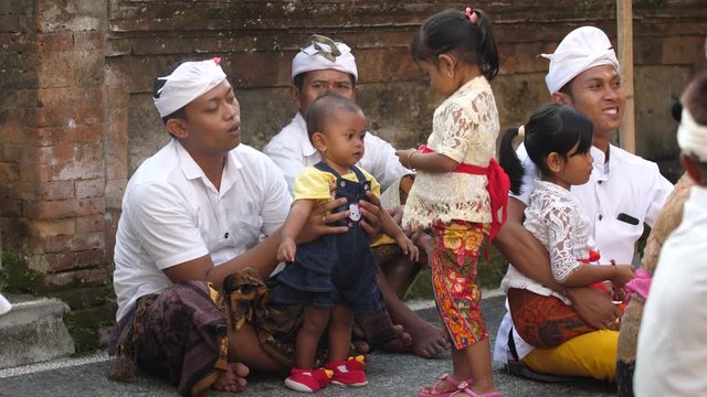 Men in traditional Balinese clothing sit on the ground outside a temple in Bali waiting for a ceremony and playing with children to entertain them. Father plays with toddler.