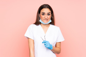 Woman dentist holding tools isolated on pink background having doubts and with confuse face...