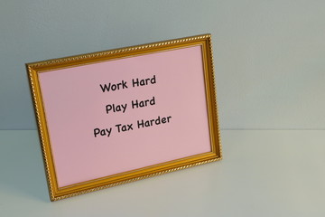 Closeup slogan Work Hard, Play Hard, Pay Tax Harder printed on pink paper in golden photo frame placed on white wooden table