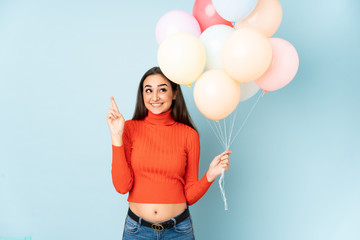 Young woman catching many balloons isolated on blue background with fingers crossing