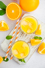 Freshly squeezed orange juice with ice in a glass with a straw on a wooden board on a light background with fresh oranges. Vertical orientation, close up, top view.