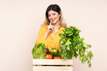 Teenager farmer girl with freshly picked vegetables in a box isolated on beige background doing silence gesture