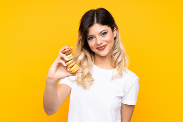 Teenager girl isolated on yellow background holding colorful French macarons and with happy expression