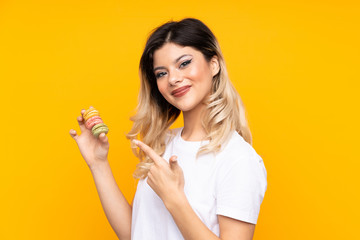 Teenager girl isolated on yellow background holding colorful French macarons and pointing it