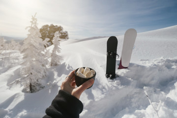 Cup of coffee with marshmallow in female hand, winter outdoor landscape, snowboards on background....