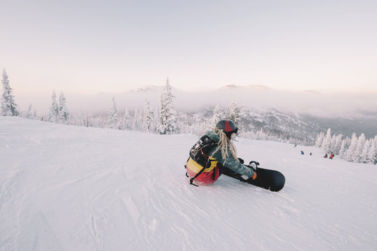 sitting female snowboarder wearing long hair preparing for riding from mountain top