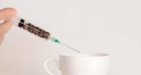 in female fingers a syringe with coffee beans over a coffee cup. At the end of the needle is a drop of coffee. Concept - Coffee Addiction