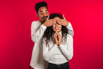 African man closes eyes of his beloved girlfriend before surprise her. Couple in white on red studio background. Love, holiday, happiness concept.