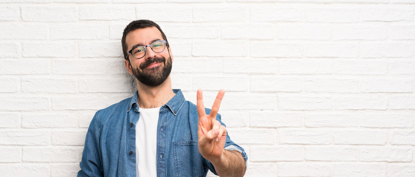 Handsome man with beard over white brick wall smiling and showing victory sign