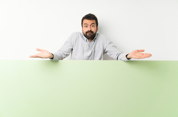 Young handsome man with beard holding a big green empty placard having doubts while raising hands