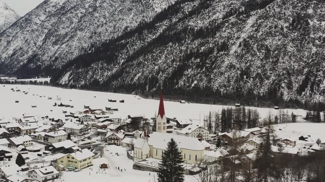 aerial view with a sideways flight behind trees to the church of holzgau austria. Beautiful view of snowy houses and church in alps. kids playing in the snow.