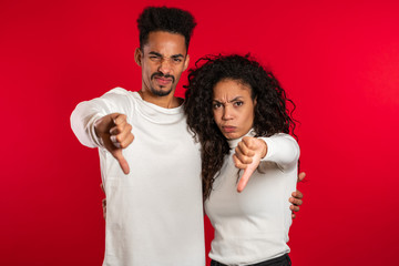 Young african couple standing on red studio background expressing discontent and showing thumb down gesture at camera. Portrait of man and woman with sign of dislike.