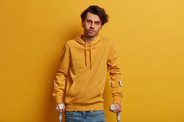 Disappointed unshaven man with deep injuries hit by car during crossing road, fell on asphalt, uses crutches because of fractures, wears casual sweatshirt, cures in clinic, has medical insurance