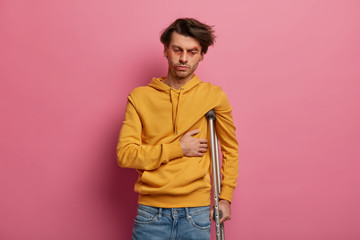 Distressed male feels pain in ribs, suffers from fracture after domestic accident, looks sadly down, thinks over treatment, poses with crutch under arm, fell while snowboarding during recreation time