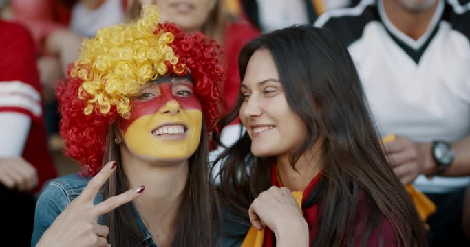 Two women sitting in fan zone with a wig and face painted in german flag colors gesturing victory sign. Devoted supporters of a Germany soccer team in stadium.
