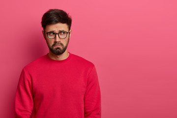Disappointed bearded man has breakup depression, looks sadly aside, wants to cry from despair, frowns face in dissatisfaction, dressed casually, isolated on pink wall, blank space on right side