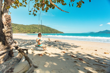 Obraz premium Beautiful girl swinging on wooden swing on the tree at the beach, happy smiling girl with red hair, playing on the beach on vacation trip, at Parnaioca beach, in Ilha Grande, in Rio de Janeiro, Brazil