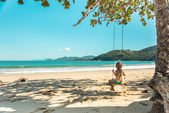 Beautiful girl swinging on wooden swing on the tree at the beach, happy smiling girl with red hair, playing on the beach on vacation trip, at Parnaioca beach, in Ilha Grande, in Rio de Janeiro, Brazil