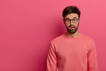 Crazy funny bearded man crosses eyes and sticks out tongue, has bored expression, foolishes around, wears spectacles and casual jumper, isolated on pink background. Postive human feelings concept