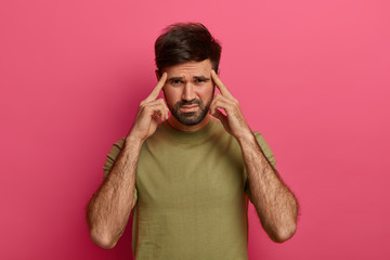 Depressed frustrated bearded man frowns and keeps index fingers on temples, cannot focus, thinks up idea, ponders on decision, feels headache or migraine, wears casual t shirt poses on pink wall