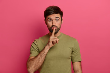 Photo of surprised bearded man tells secret, keeps index finger on lips, makes shush or shh...