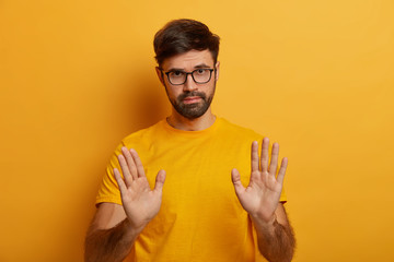 Slow down. Serious looking displeased bearded man shows stop gesture, asks to hold horses, keeps palms towards camera, says take it easy and control your behaviour, stands against yellow background - obrazy, fototapety, plakaty