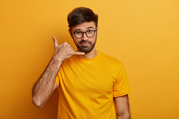 Confident charismatic man makes call me gesture, communicates with gestures, asks girlfriend telephone number, looks with flirty expression, isolated on yellow wall, wears casual outfit. Body language - obrazy, fototapety, plakaty