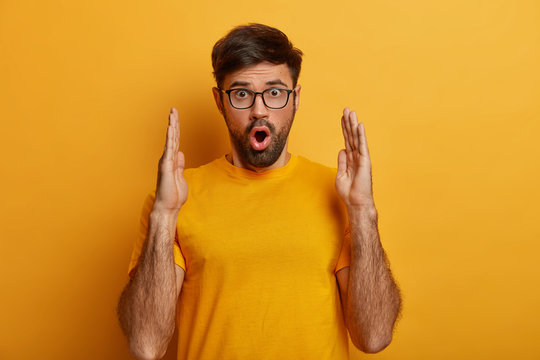 Shocked embarrassed young man demonstrates something very huge, gasps from wonder, measures big object with both hands, wears casual yellow t shirt, stands indoor, keeps mouth opened, says omg