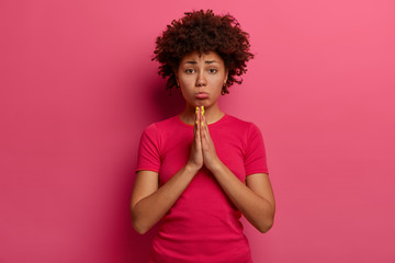 Frustrated unhappy curly haired woman begs for permission, keeps palms pressed together, purses...