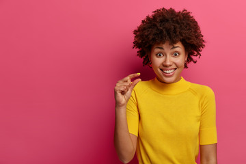 Studio shot of curly haired positive woman measures tiny object, shows something very little, has glad expression, wears yellow t shirt, talks about size, isolated on pink wall, copy space aside - obrazy, fototapety, plakaty