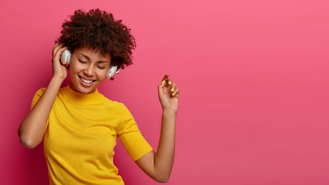 Optimistic carefree woman with glad expression dances carefree, keeps arm raised in air, moves with rhythm of music, wears headphones on ears, closes eyes, has upbeat mood, poses over rosy background