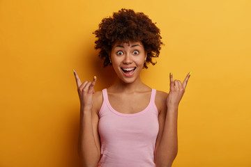 Photo of surprised African American woman makes rock n roll hand sign, enjoys listening favourite...
