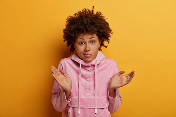 Indecisive curly woman raises palms in clueless gesture, looks doubtfully, faces difficult choice, wears casual anorak, has doubts, considers if she did right, has questioned expression, poses indoor © WHstudio Leushin N