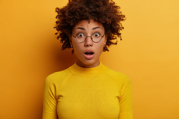 Fototapeta na wymiar Concerned shocked African American woman opens mouth, witnesses shocking scene, has bugged eyes, wears spectacles and yellow sweater, reacts to fresh amazing gossips, poses over bright wall.