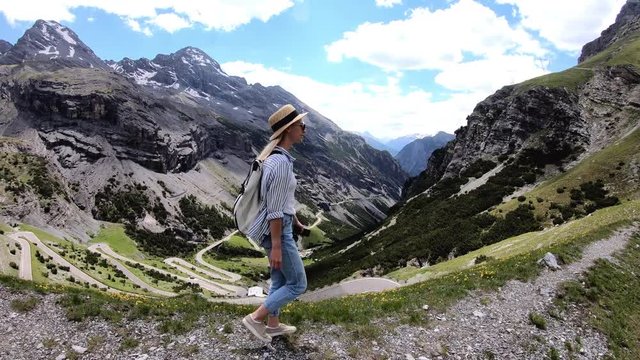Positive caucasian young woman in hat walking on mountain road viewpoint of Stelvio Hairpins Pass exploring scenic landscape and nature, female tourist with backpack enjoying recreation time on path