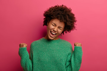 Euphoric curly haired woman winner raises hands with clenched fists, makes yes gesture, celebrates importnat achievement or success, wears green sweater, got prize, isolated on pink background