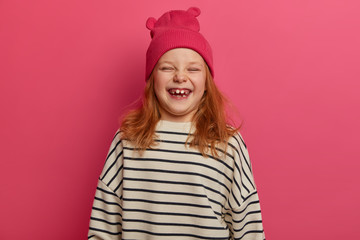 Children and happiness concept. Joyful redhead girl laughs out from something funny, wears pink hat with ears and loose striped sweater, smiles brightfully, has missing teeth, models indoor.