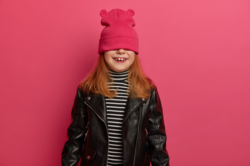 Studio shot of lovely redhead girl plays hide and seek game, waits for surprise with positive emotions, covers eyes with pink hat, wears striped jumper and leather jacket, has fun, poses indoor