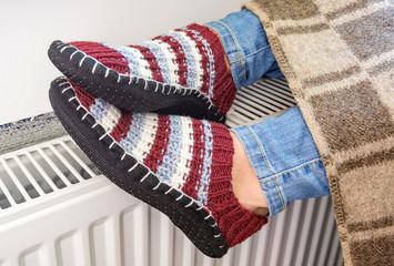 A woman is dressed in warm knitted woolen slippers and heats her legs on a heating radiator at home.