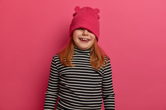 Waist up shot of adorable girl has playful mood, looks from above hat, foolishes around, wears striped poloneck, has broad smille, poses against rosy background, being disobedient or naughty