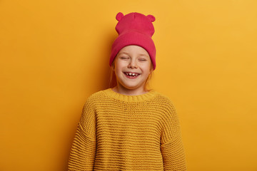 Little cute girl laughs happily, feels glad to receive new toy doll from parents, closes eyes, has fun indoor, wears hat with ears and loose knitted sweater, isolated over yellow background.