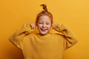 Horizontal shot of optimistic merry small kid plugs ears with index fingers, chuckles positively,...
