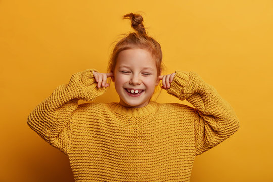 Horizontal shot of optimistic merry small kid plugs ears with index fingers, chuckles positively, has ginger hair bun, wears oversized knitted sweater, isolated on yellow background. Stop this sound