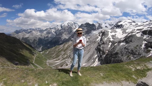 Happy caucasian hipster girl standing on breathtaking mountains background making selfie on smartphone camera, young female wanderlust blogger shooting video for travel blog during summer vacations
