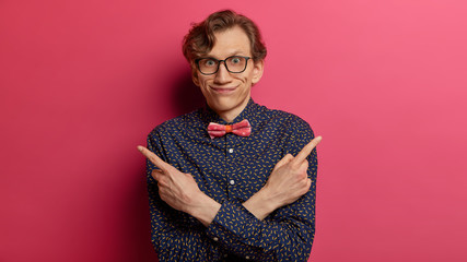 Confused male nerd wears elegant shirt with bowtie, points sideways, tries to choose between two...