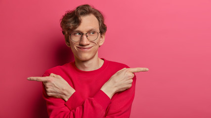 Mysterious delighted man ponts to both sides with index fingers, shows different directions, purses lips and looks aside, offers two options for choosing, wears transparent glasses and red jumper